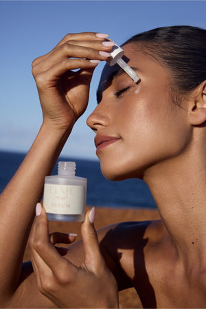 Woman applying serum with a dropper bottle against a blue sky and ocean background